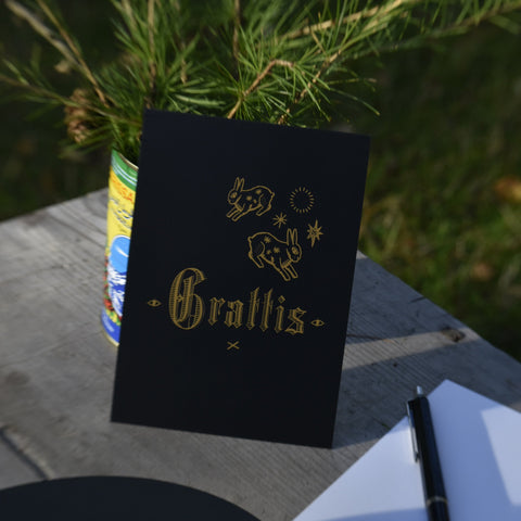 A black Grattis (congrats) greeting card with rabbit illustrations, displayed on a rustic wooden table next to a pen and white card back, with green foliage in the background.