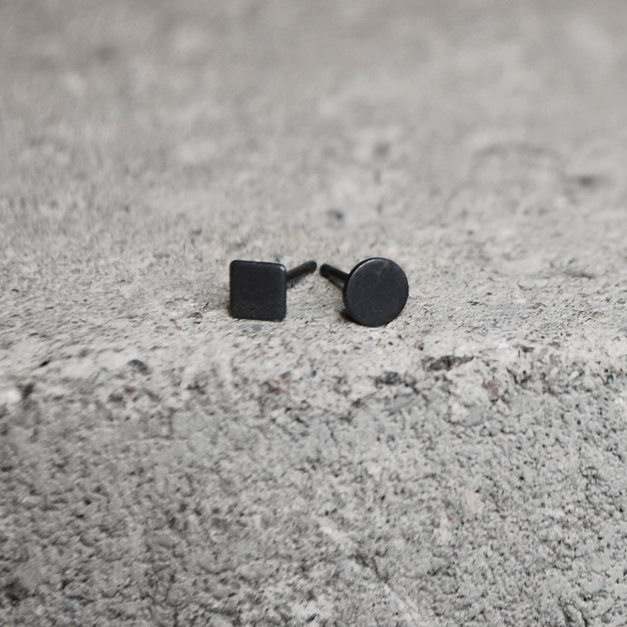 A pair of matte black stud earrings, one with a square face and one with a round face, resting on a textured gray concrete surface.