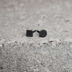 A pair of matte black stud earrings, one with a square face and one with a round face, resting on a textured gray concrete surface.