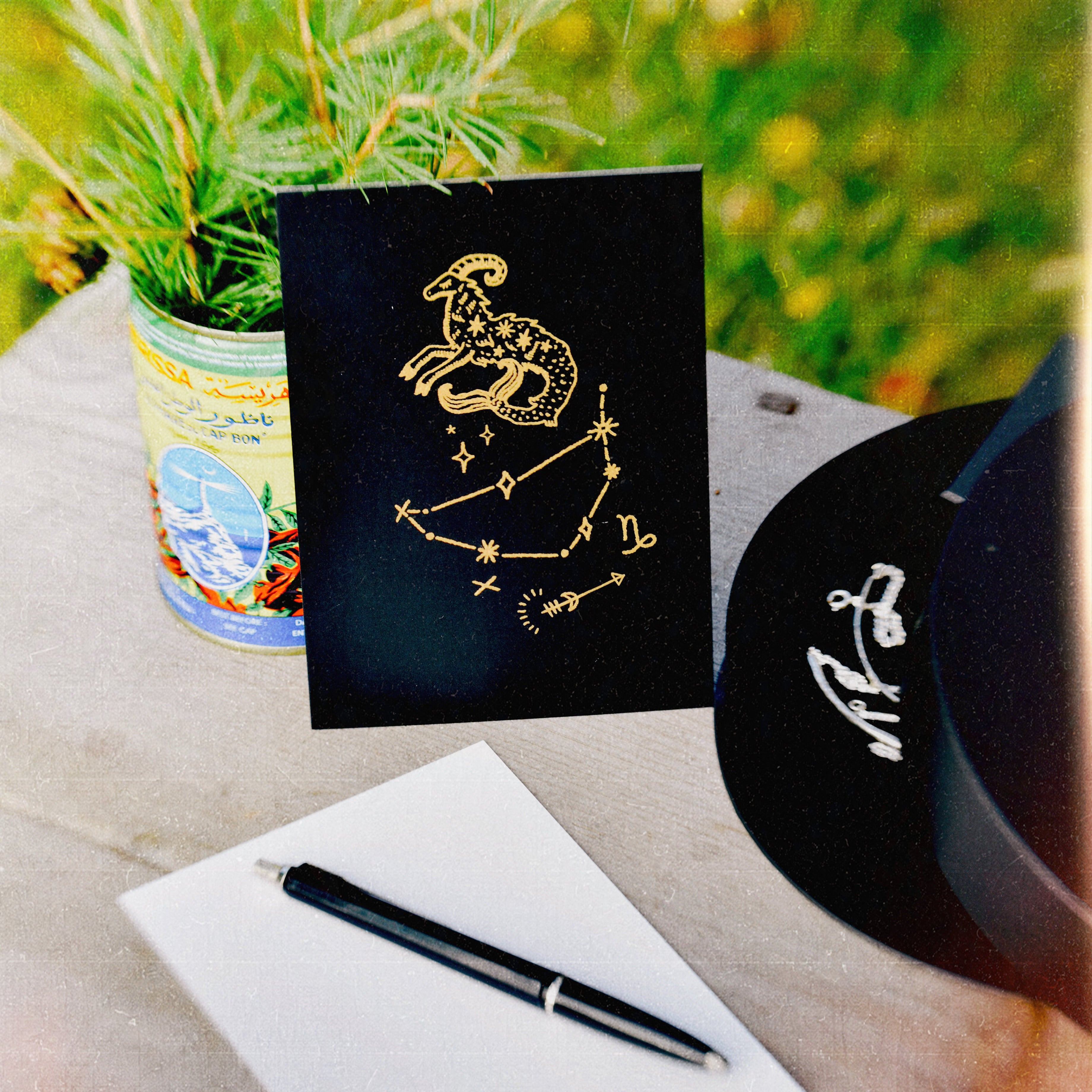 A matte black card with a zodiac illustration constellation design of Capricorn (stenbocken in Swedish), styled on a rustic wooden table with a white card backside, a black pen, and a plant in a blurred outdoor setting.